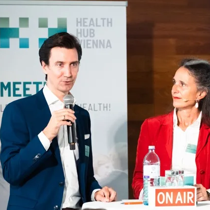 Health Hub Vienna A woman in a red jacket watches a man talking into a microphone