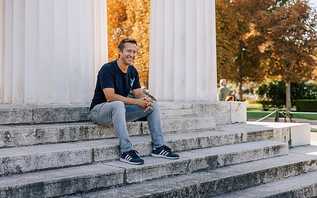 Man in a black tshirt and jeans sitting on building steps in a park in autumn
