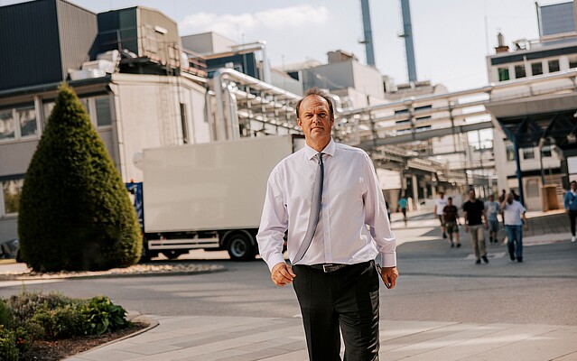 Smiling man in a white shirt with blue tie walking towards the camera across a street