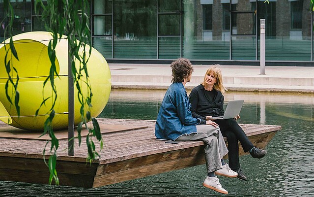 Two women discussing on a pier among greenery.