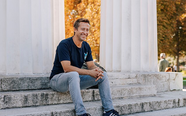 Man in a black tshirt and jeans sitting on building steps in a park in autumn