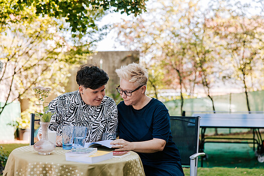 Two women sitting at a table, reading a book together.