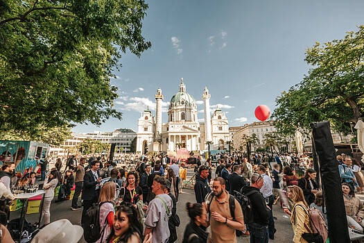 A crowd of ViennaUP participants chat at the Homebase, with the majestic St. Charles Church lit by sunlight and rising in the background.