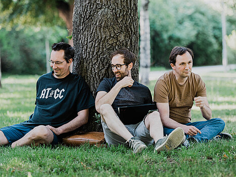 Three men sitting in the grass next to a tree