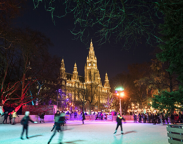 Ice skaters on an ice rink in front of a large, illuminated building