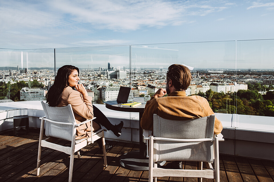 Woman and man sitting and speaking on a rooftop Vienna cityscape in front of them