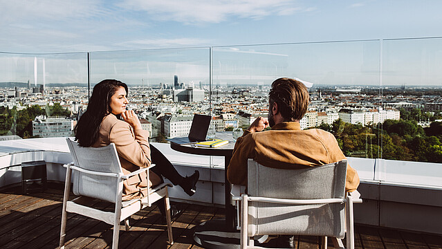 Woman and man sitting and speaking on a rooftop Vienna cityscape in front of them