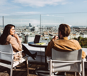 Woman and man sitting and speaking on a rooftop Vienna cityscape in front of them