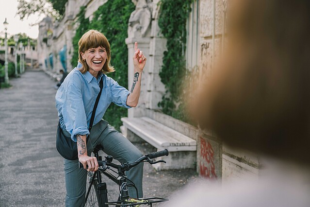 A young female startup executive is shown riding her bicycle in Vienna's Stadtpartk and waving to a friend.