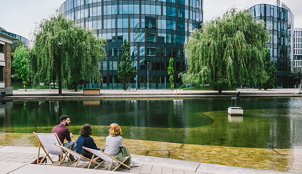 Two women and a man sitting in front of a lake and modern business buildings in Vienna